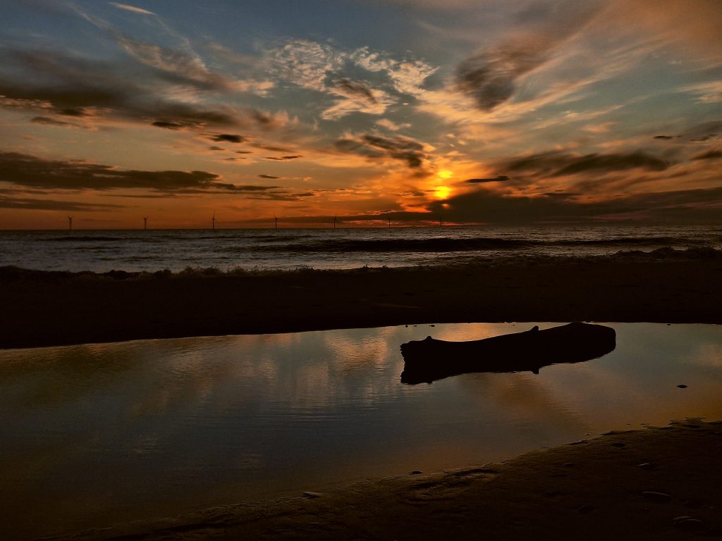 Sonnenuntergang am Strand. Im Vordergrund ein Baumstamm im Wasser.